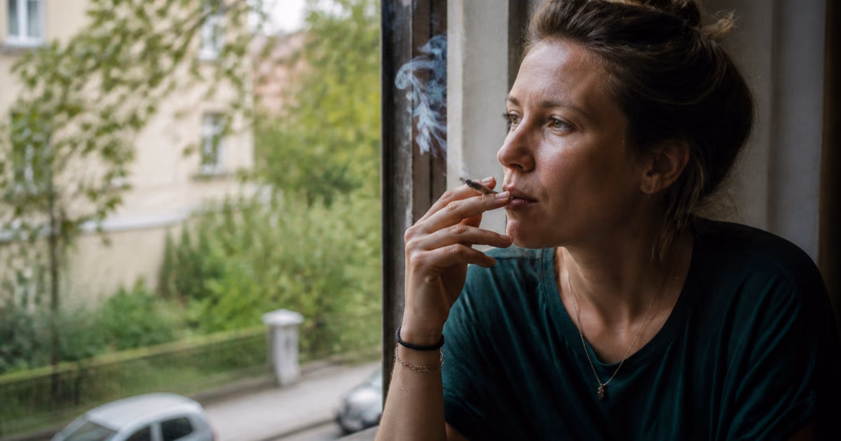 Woman smoking a joint by a window, looking out onto a leafy street